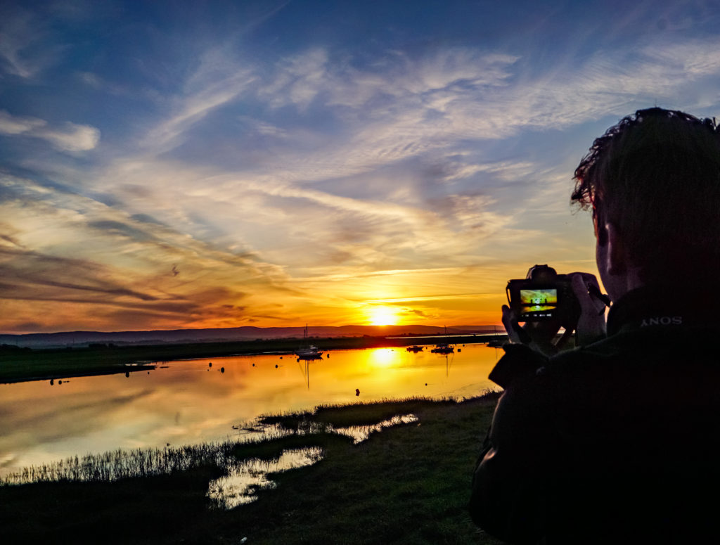 A man takes a picture of a bay at sunset