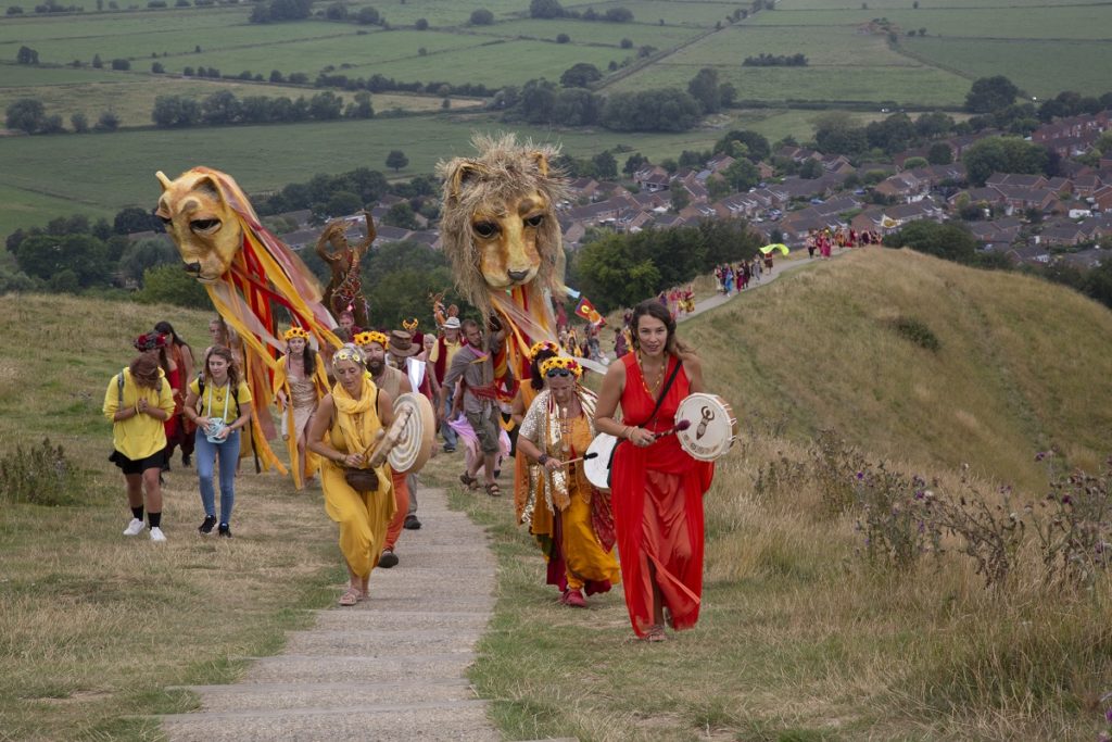 People in bright clothes march up Glastonbury Tor with drums and lion puppets