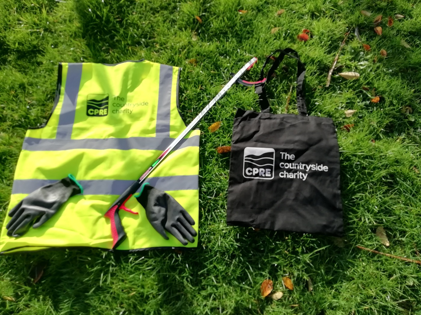 A hi-vis vest, a litterpicking stick, a pair of gloves and a branded bag laid out on grass